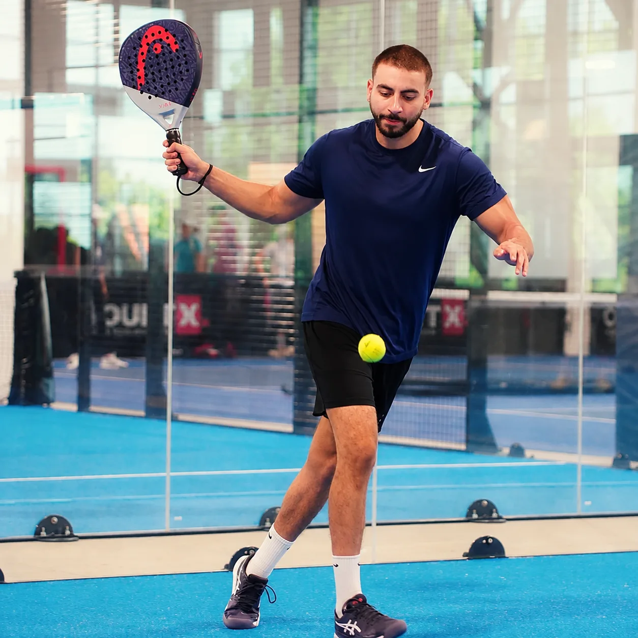 Setting up a padel shot with the ball in the air, glass-walled court behind