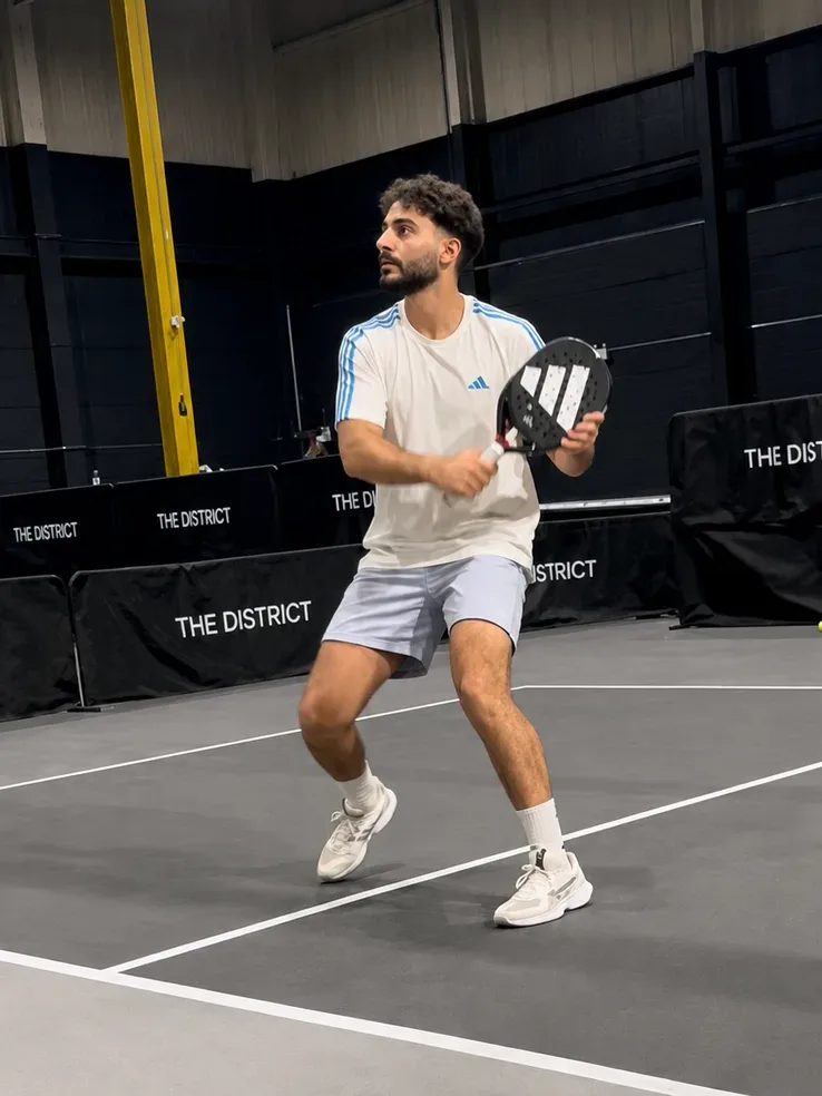 Waiting on the return at an indoor padel court, boards around the court reading The District