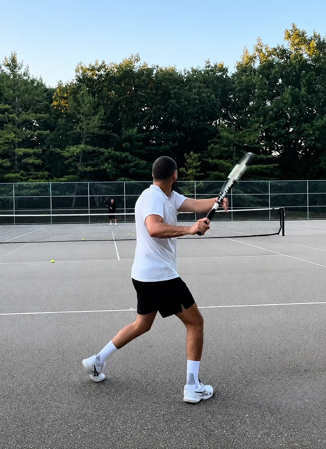 Winding up a tennis backhand on an outdoor court at dusk, treeline behind
